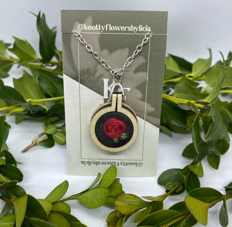 hand stitched Red woven rose with multiple shades of light, medium, and dark red around and two small green leaves on a black background. the pendant is on a silver chain