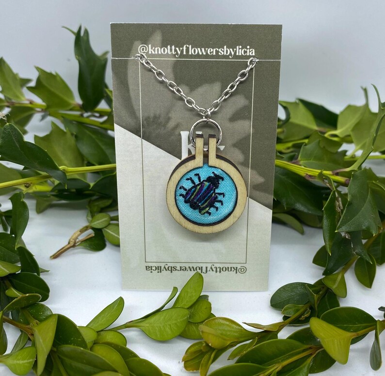 Hand stitched embroidered beetle bug pendant with purple, blue, and green vertical stripes on the bug&#39;s back and 4 legs on each side.It has large antennae on its head. The bug is on a sky blue background and on a silver chain