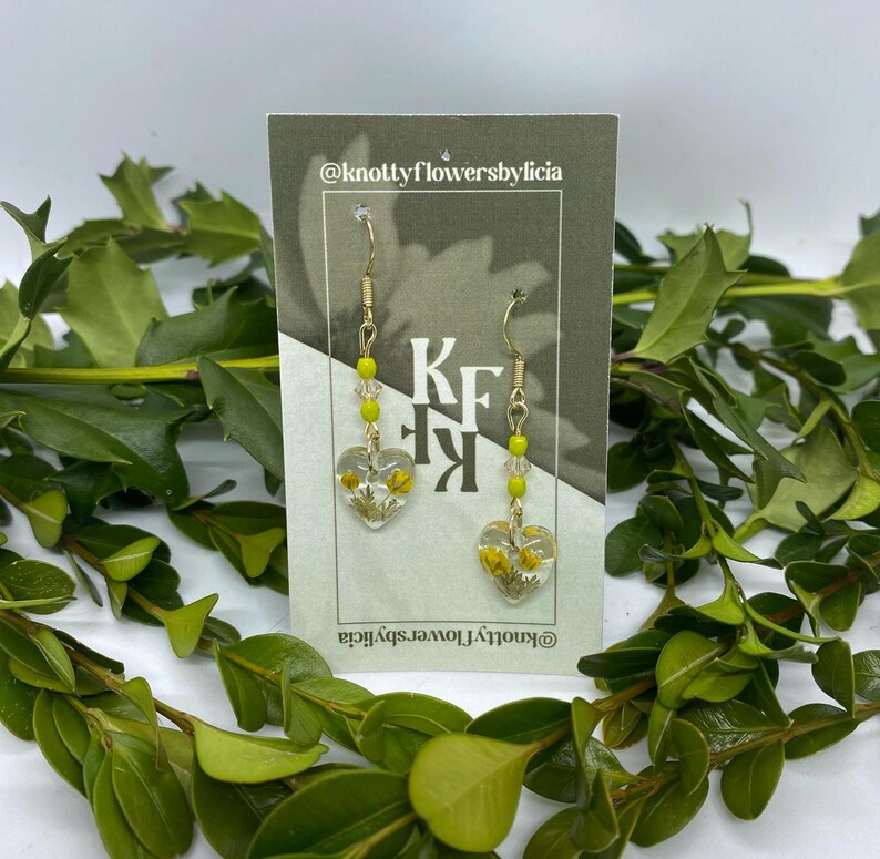 Handmade resin heart shaped earrings with tiny yellow flowers and super small green leaves and small green ceramic beads with faintly golden yellow glass beads in the middle